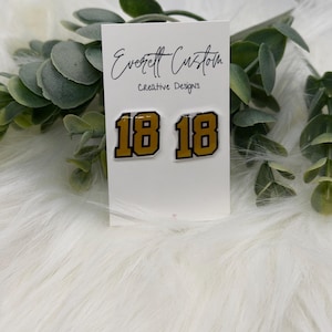 May include: A pair of gold and black number 18 earrings on a white card with the text "Everett Custom Creative Designs". The earrings are displayed on a white fluffy surface with green foliage.