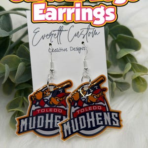 May include: Custom logo earrings featuring the Toledo Mud Hens baseball team logo. The earrings are made of a clear material with a silver hook. The logo is in orange, black, and white. The text "Custom Logo Earrings" is at the top.