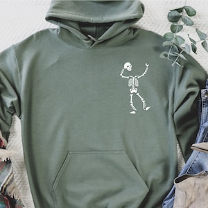 K&ouml;nnte beinhalten: Salbeigr&uuml;ner Kapuzenpullover mit wei&szlig;em Skelett-Motiv. Der Hoodie hat eine K&auml;ngurutasche und eine Kapuze mit Kordelzug. Ein karierter Schal, blaue Jeans und beige Stiefel sind um den Hoodie angeordnet.