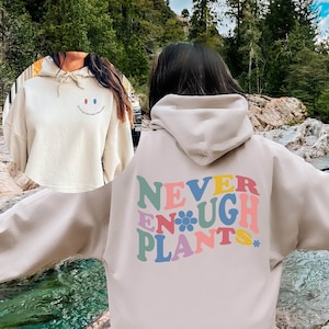May include: A beige oversized hoodie with a colorful graphic on the back that says "NEVER ENOUGH PLANTS" with a flower in the middle of the word "ENOUGH".