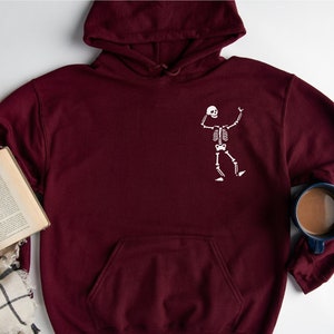 K&ouml;nnte beinhalten: Ein weinroter Kapuzenpullover mit einem wei&szlig;en Skelett-Motiv auf der Vorderseite. Der Hoodie hat eine K&auml;ngurutasche und eine Kapuze mit Kordelzug. Ein Buch, eine Brille und ein Schal liegen neben dem Hoodie.