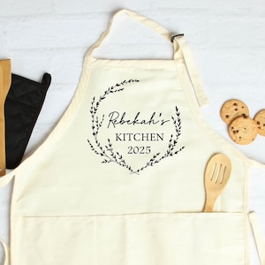 K&ouml;nnte beinhalten: Eine wei&szlig;e Baumwollsch&uuml;rze mit einer gro&szlig;en Tasche und verstellbaren Tr&auml;gern. Die Sch&uuml;rze hat einen schwarzen Aufdruck einer Blumengirlande mit dem Text "Rebekah's Kitchen 2025" darin.