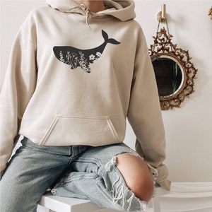 May include: A beige hoodie with a black silhouette of a whale with floral details inside the whale's body. The model is wearing the hoodie and blue jeans.