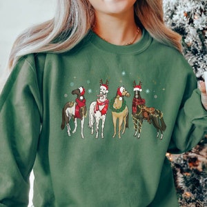 May include: Green sweatshirt featuring a festive graphic of five horses adorned with Christmas decorations. Each horse wears a Santa hat, and some have scarves and decorative lights. Snowflakes are scattered around the horses, creating a holiday theme.
