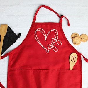 May include: Red kitchen apron with a white heart and the word "Gigi" inside. The apron has a large pocket on the front and two ties.