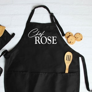 May include: Black apron with the text "Chef ROSE" in white script. The apron has two pockets and is paired with a black oven mitt, a wooden spoon, and chocolate chip cookies.