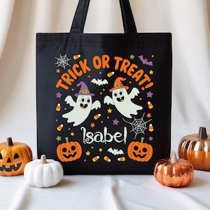 May include: Black Halloween tote bag with the words "Trick or Treat!" and the name "Isabel". The bag features two cartoon ghosts wearing witch and wizard hats, surrounded by pumpkins, bats, and candy corn.