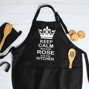 May include: Black kitchen apron with a white crown and the text "Keep Calm Because Rose Is In The Kitchen".