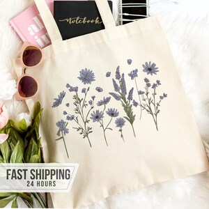 May include: A cream-colored tote bag with a floral design featuring blue and purple flowers. The bag is filled with a pink notebook, a black notebook with "Notebook" in gold lettering, a smartphone, and sunglasses. The words "FAST SHIPPING 24 HOURS" are printed on a white banner.