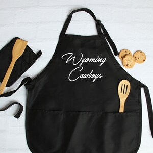 May include: Black kitchen apron with two pockets and a white embroidered design that says "Wyoming Cowboys". The apron is shown with a wooden spoon and a pot holder.