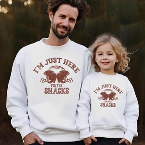 May include: White sweatshirts with a brown graphic print of two football helmets and the text "I'm just here for the snacks".