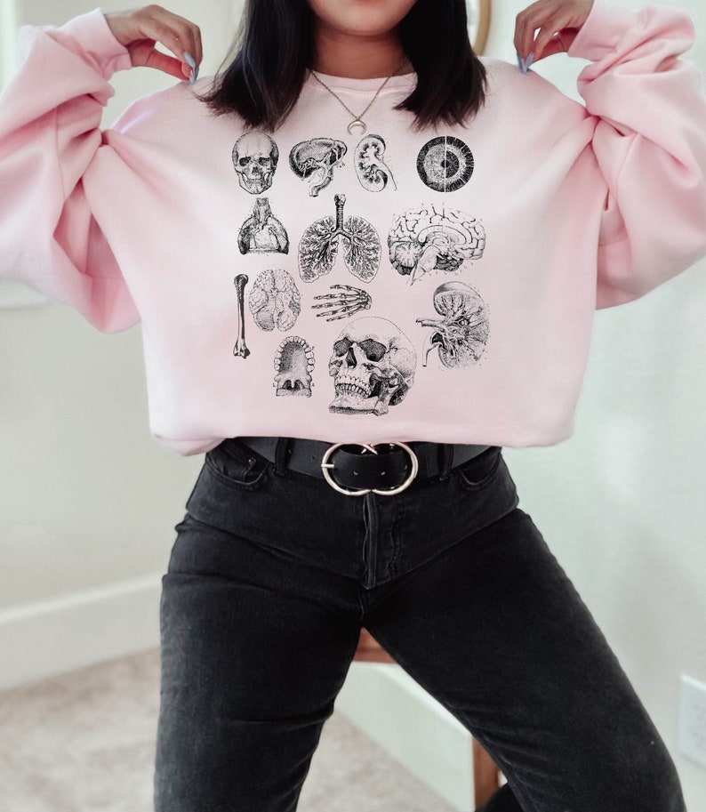 May include: A pink sweatshirt with a vintage anatomical illustration of a skull, brain, lungs, heart, and other organs. The sweatshirt is worn by a person with black jeans and a silver belt.