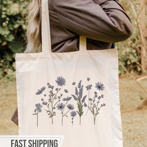 May include: Cream-colored tote bag featuring a floral design with blue and green accents. The bag has a rectangular shape with two long straps. The text "FAST SHIPPING 24 HOURS" is displayed on the bottom.
