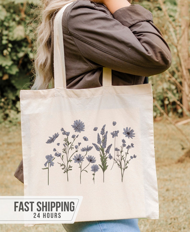 May include: Cream-colored tote bag featuring a floral design with blue and green accents. The bag has a rectangular shape with two long straps. The text "FAST SHIPPING 24 HOURS" is displayed on the bottom.