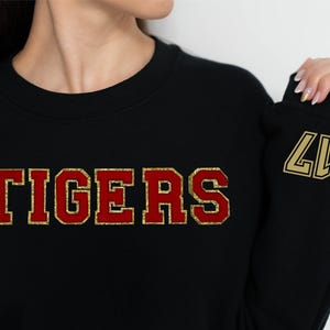 May include: A black crewneck sweatshirt featuring the word TIGERS in large, red, glittery block letters with a gold outline across the chest. The number 17 is printed in gold on the left sleeve near the cuff in a bold, outlined font style.