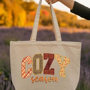 A hand holding a beige tote bag with the word "COZY" printed on it, set against a background of a field with autumn foliage.