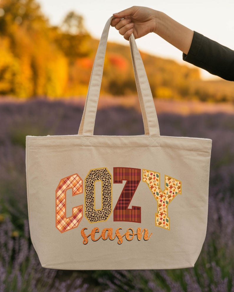 A hand holding a beige tote bag with the word "COZY" printed on it, set against a background of a field with autumn foliage.
