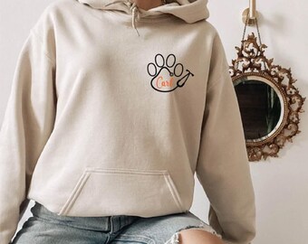 May include: A beige hooded sweatshirt featuring a black graphic on the left chest. The graphic depicts a paw print combined with a stethoscope, with the name Carl written in orange script inside the paw pad area. The hoodie has a front pocket.