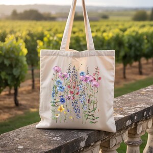 A white tote bag with a floral design printed on it, hanging on a stone railing in a vineyard.