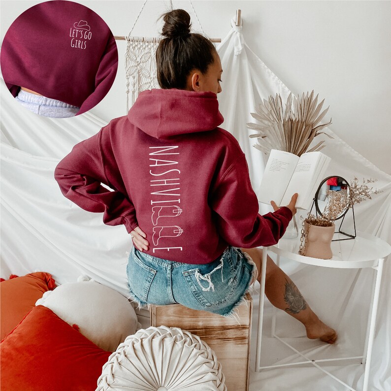 May include: A maroon cropped hoodie with a white graphic that says "Nashville" with a pair of boots on the side. The model is wearing denim shorts and has a tattoo on their arm.