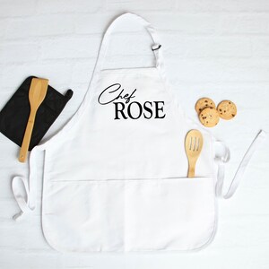 May include: White apron with the text "Chef ROSE" in black script. A wooden spatula and slotted spoon are in the pockets. A black oven mitt and three chocolate chip cookies are also present.