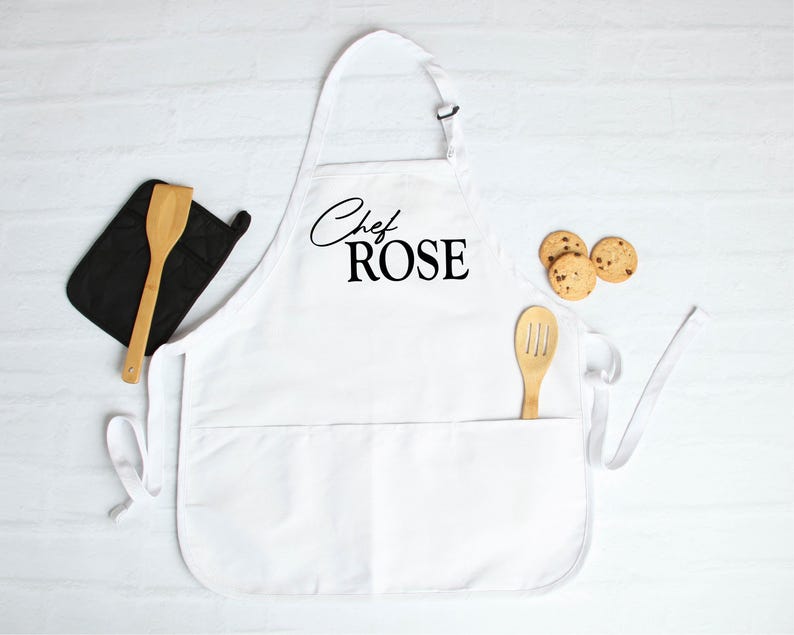 May include: White apron with the text "Chef ROSE" in black script. A wooden spatula and slotted spoon are in the pockets. A black oven mitt and three chocolate chip cookies are also present.