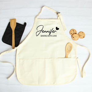 Peut inclure: Tablier cr&egrave;me avec le nom "Jennifer" et la phrase "BAKING WITH LOVE" en &eacute;criture noire. Une cuill&egrave;re en bois est dans la poche, et une manique noire et trois biscuits aux p&eacute;pites de chocolat sont &agrave; proximit&eacute;. Le tablier a une sangle de cou et des liens &agrave; la taille.