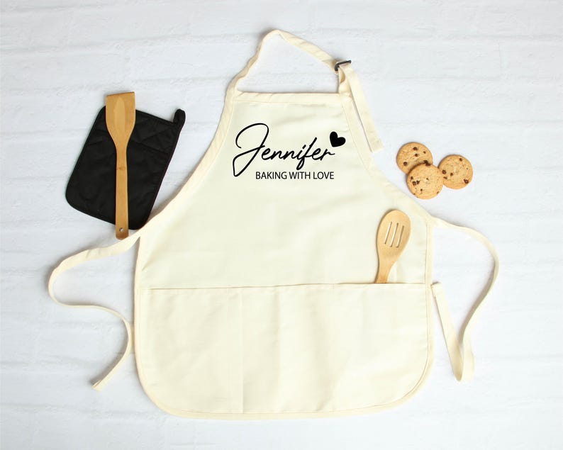 Peut inclure: Tablier cr&egrave;me avec le nom "Jennifer" et la phrase "BAKING WITH LOVE" en &eacute;criture noire. Une cuill&egrave;re en bois est dans la poche, et une manique noire et trois biscuits aux p&eacute;pites de chocolat sont &agrave; proximit&eacute;. Le tablier a une sangle de cou et des liens &agrave; la taille.