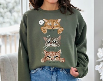 May include: An olive green crewneck sweatshirt featuring a graphic of three cats stacked vertically. The top cat is orange and labeled RELAX, the middle cat is grey tabby and labeled CURIOUS, and the bottom cat is brown tabby and labeled CAUTIOUS.