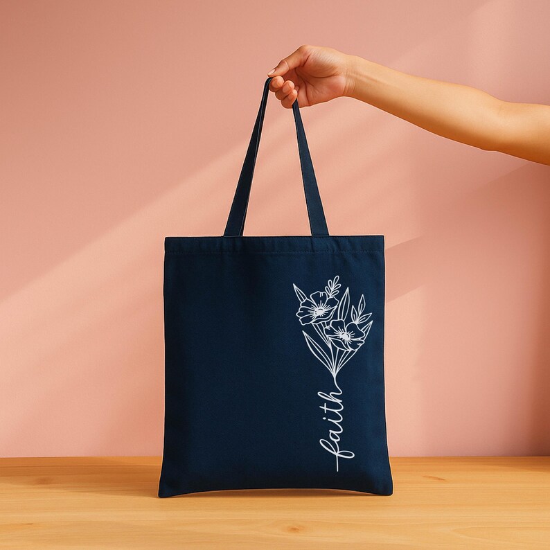 A navy blue tote bag with a white floral design and the word "faith" printed on it, held by a hand against a pink background.