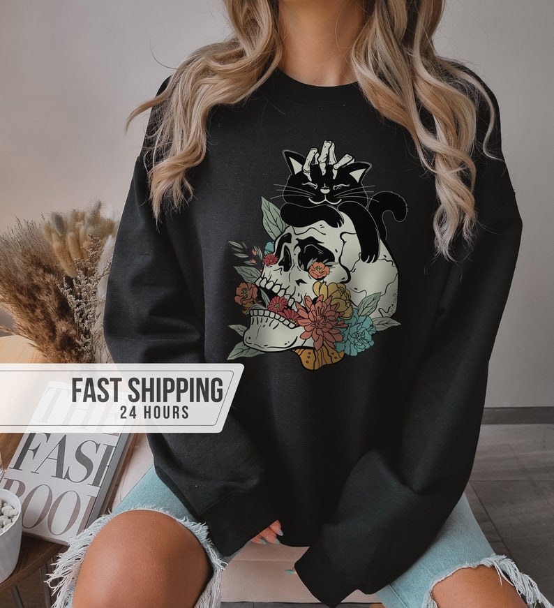 May include: Black sweatshirt featuring a skull with floral accents and a black cat. The skull has colorful flowers and leaves around it. The cat is sitting on top of the skull. The text "FAST SHIPPING 24 HOURS" is on the left side of the image.
