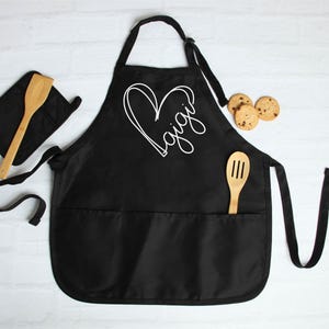 May include: Black apron with a large white heart and the word "Gigi" inside. The apron has a large pocket and two smaller pockets on the sides. It comes with a matching black pot holder and a wooden spoon.