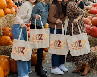 May include: Four beige tote bags, each bearing the words "COZY season" in patterned letters. The bags are held by people, with pumpkins and hay bales in the background, evoking an autumnal atmosphere.