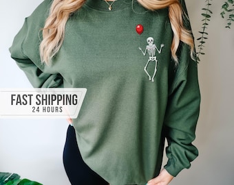May include: An olive green crewneck sweatshirt featuring a small graphic on the upper left chest of a white skeleton holding a single red balloon. The long-sleeved pullover has a ribbed neckline, cuffs, and hem, providing a simple, solid-colored design.