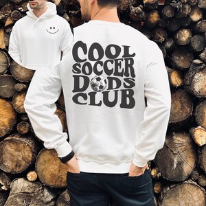 Pu&ograve; includere: Felpa bianca con una stampa grafica nera che dice "COOL SOCCER DADS CLUB" con un pallone da calcio al centro del testo.