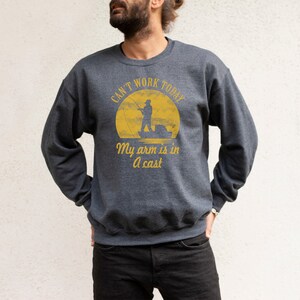 May include: A dark gray sweatshirt with a yellow graphic of a fisherman in a boat. The text on the graphic reads "Can't work today, My arm is in a cast."
