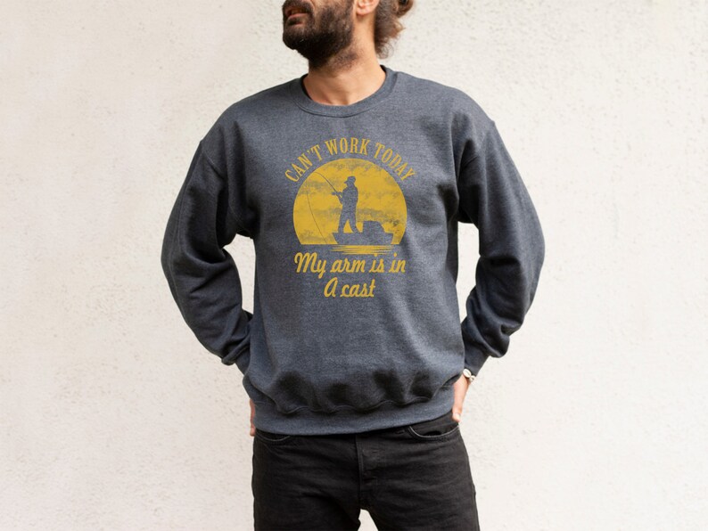 May include: A dark gray sweatshirt with a yellow graphic of a fisherman in a boat. The text on the graphic reads "Can't work today, My arm is in a cast."