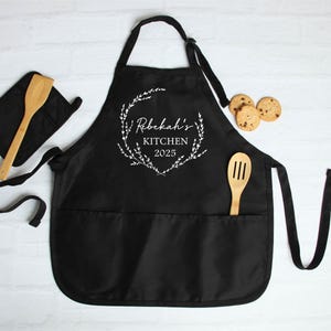 K&ouml;nnte beinhalten: Schwarze K&uuml;chensch&uuml;rze mit einem wei&szlig;en floralen Kranzdesign und dem Text "Rebekah's Kitchen 2025". Die Sch&uuml;rze hat zwei gro&szlig;e Taschen und einen passenden Topflappen.