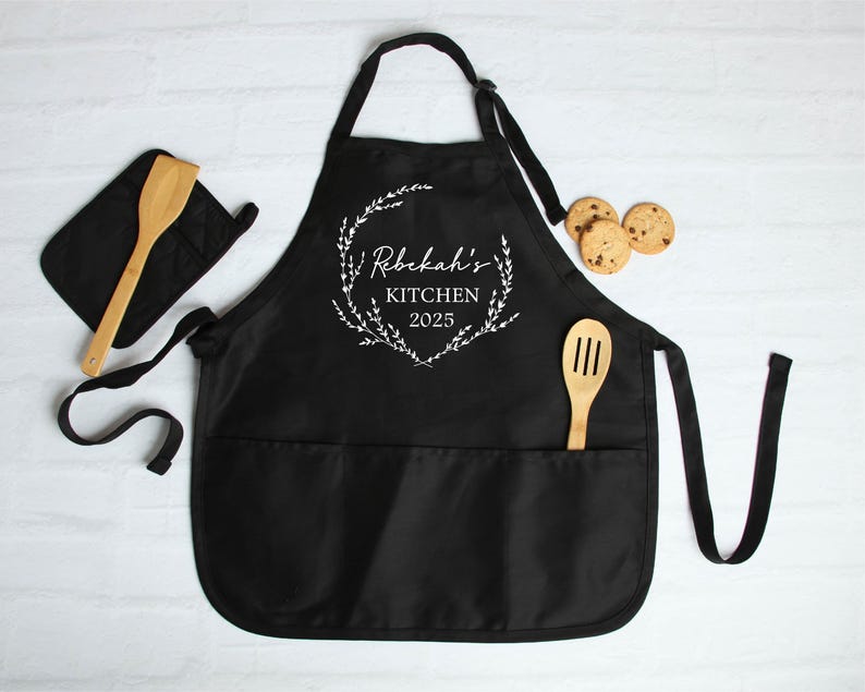 K&ouml;nnte beinhalten: Schwarze K&uuml;chensch&uuml;rze mit einem wei&szlig;en floralen Kranzdesign und dem Text "Rebekah's Kitchen 2025". Die Sch&uuml;rze hat zwei gro&szlig;e Taschen und einen passenden Topflappen.