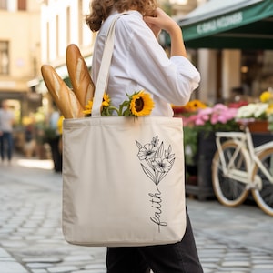A person carrying a white tote bag with a floral design and the word "faith" printed on it, along with a bouquet of sunflowers.