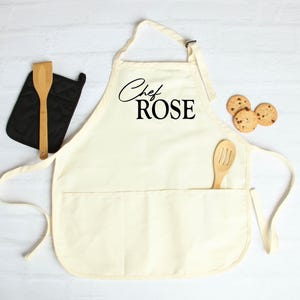 May include: Cream-colored apron with the text "Chef ROSE" in black script. A wooden spoon is in the pocket, and a black oven mitt and wooden spatula are to the left. Three chocolate chip cookies are on the right.