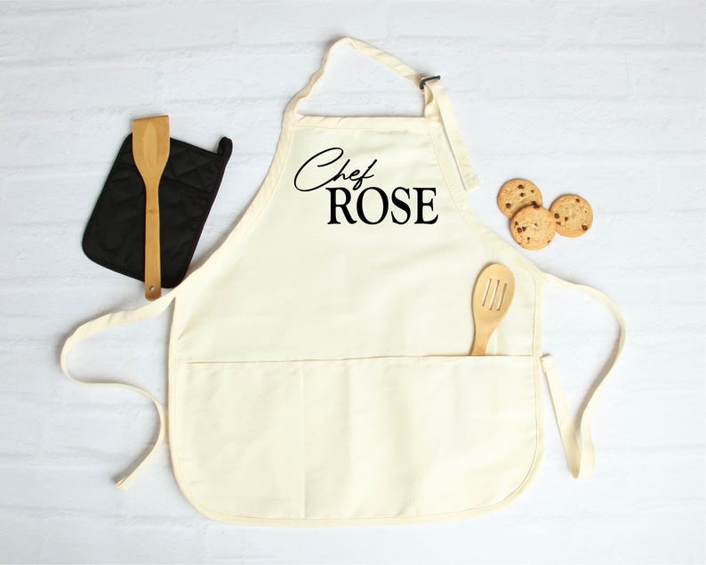 May include: Cream-colored apron with the text "Chef ROSE" in black script. A wooden spoon is in the pocket, and a black oven mitt and wooden spatula are to the left. Three chocolate chip cookies are on the right.