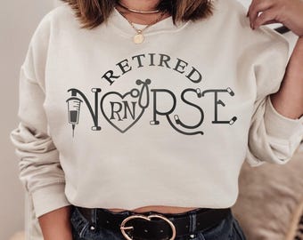 May include: A beige crewneck sweatshirt featuring dark gray text that reads RETIRED NURSE. The letter U is replaced by a heart shape containing the letters RN, and the letter N is stylized as a syringe, while the letter R incorporates a stethoscope.