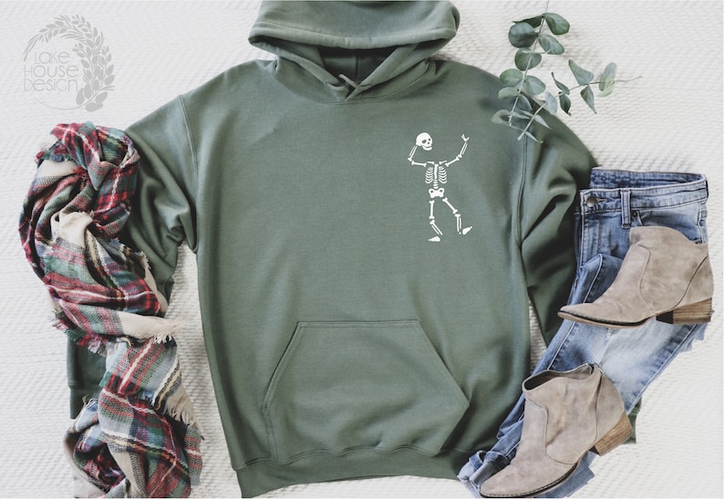 K&ouml;nnte beinhalten: Salbeigr&uuml;ner Kapuzenpullover mit wei&szlig;em Skelett-Motiv. Der Hoodie hat eine K&auml;ngurutasche und eine Kapuze mit Kordelzug. Ein karierter Schal, blaue Jeans und beige Stiefel sind um den Hoodie angeordnet.