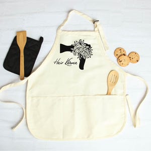 May include: A white cotton apron with a black graphic of a hairdryer with flowers and the text "Hair Karma". The apron has a large pocket and two smaller pockets.