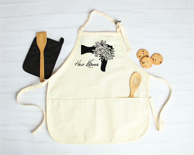 May include: A white cotton apron with a black graphic of a hairdryer with flowers and the text "Hair Karma". The apron has a large pocket and two smaller pockets.