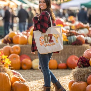 A woman stands in front of a display of pumpkins and hay bales, holding a bag with the word "COZY" printed on it.