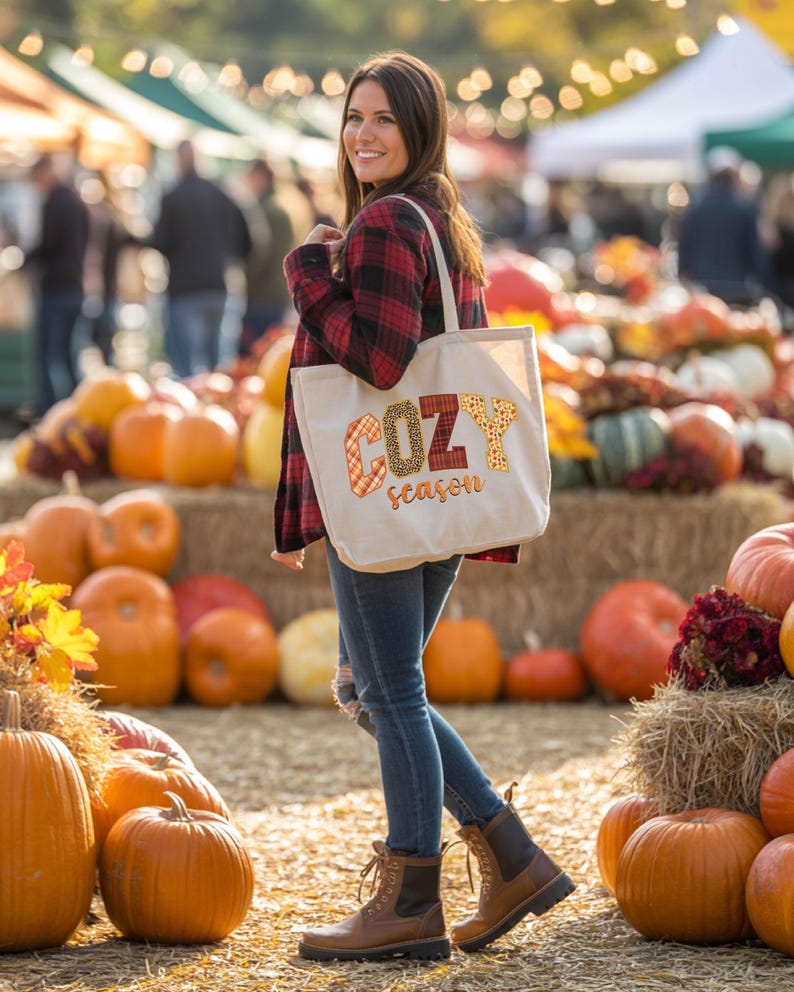 A woman stands in front of a display of pumpkins and hay bales, holding a bag with the word "COZY" printed on it.