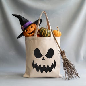 A Halloween-themed tote bag with a carved jack-o-lantern and a broom, along with a small pumpkin and gourds, are displayed against a gray background.