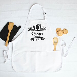 May include: White apron with a black design that says "Nana's Garden" with gardening tools and flowers. The apron has a large pocket in the front.